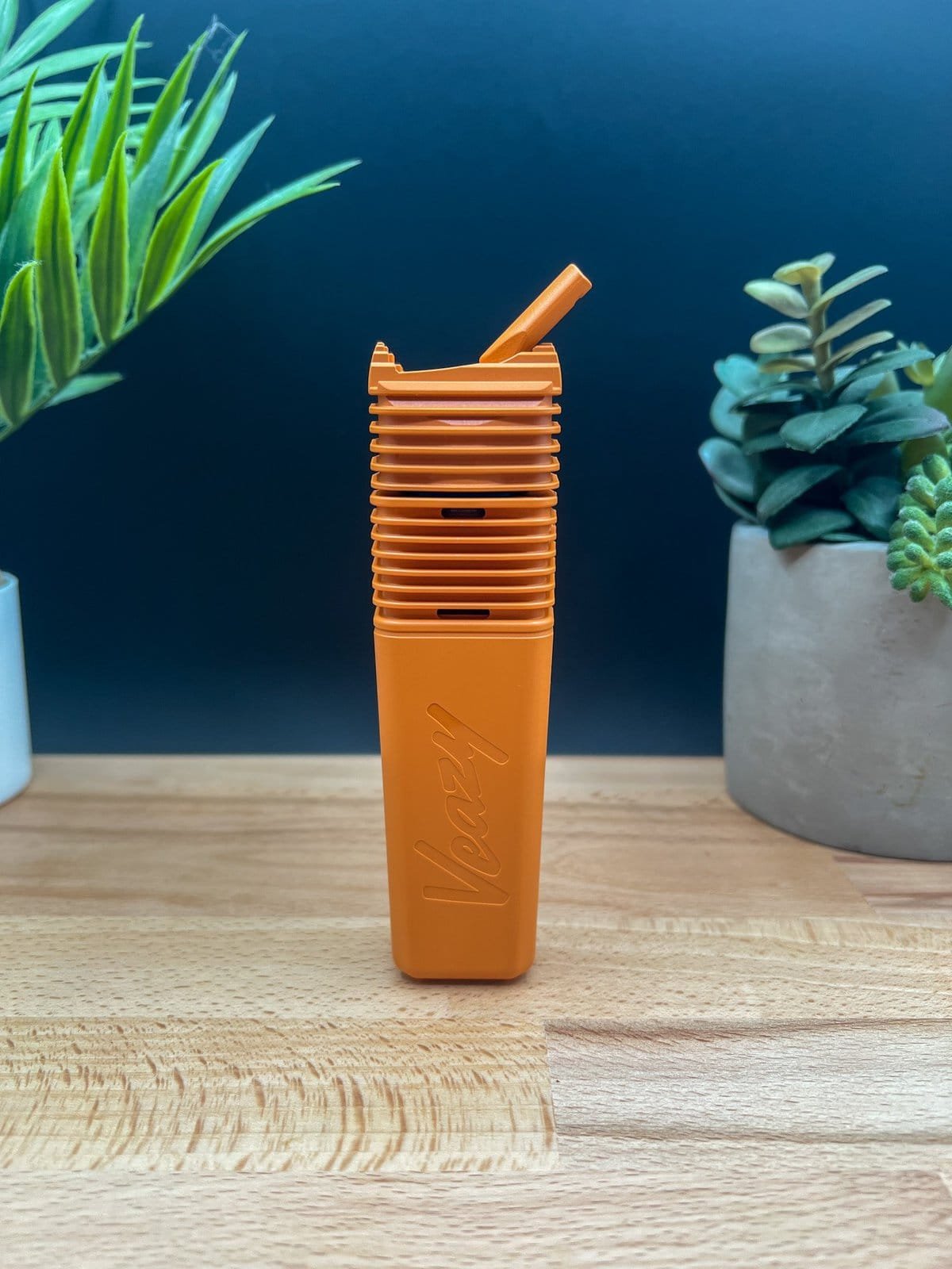 Orange Storz & Bickel Veazy vaporizer standing upright on a wooden surface with plants in the background — featured in our Veazy Review.